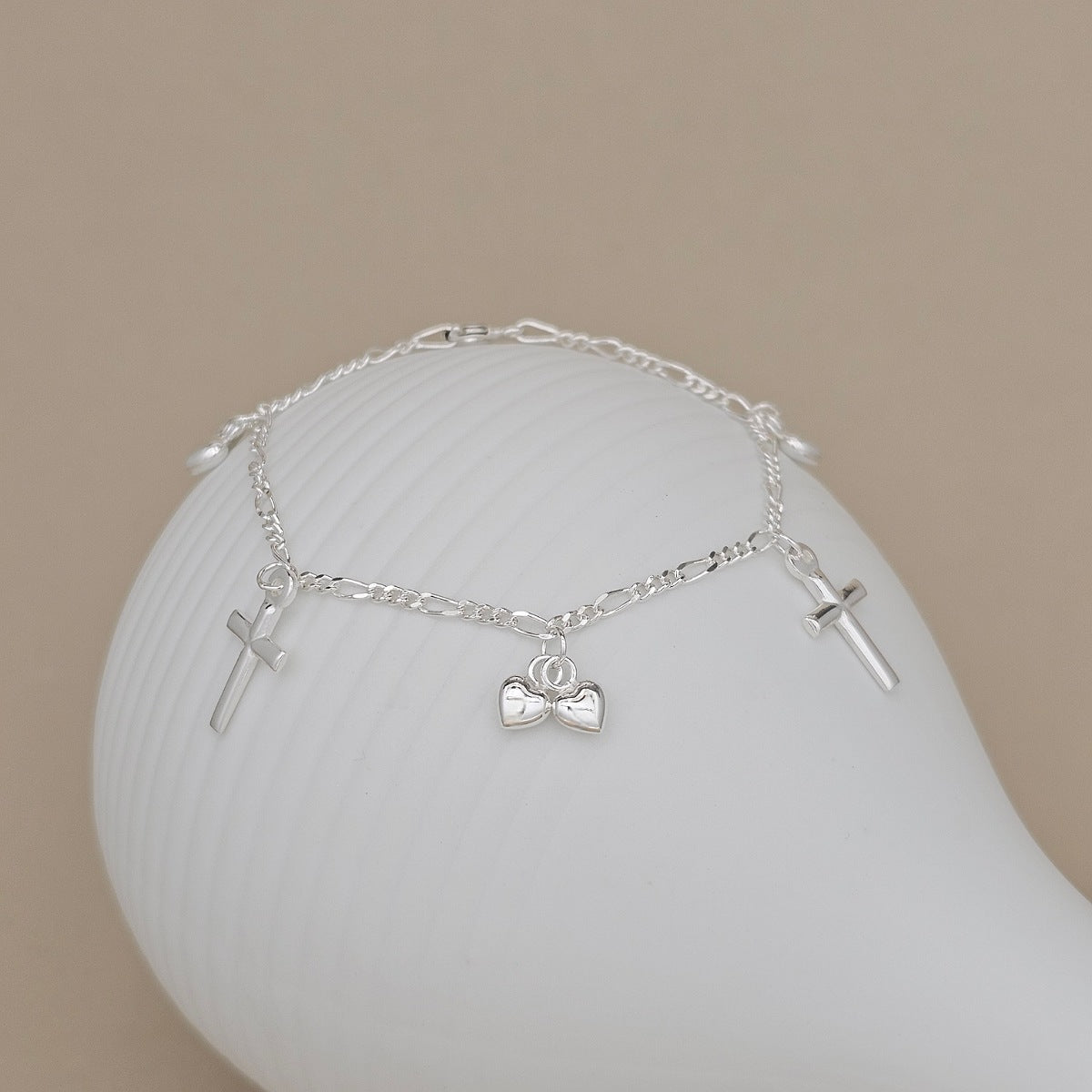 A delicate silver chain bracelet with three charms, including two small crosses and heart charms, displayed on a white curved stand against a warm beige background.