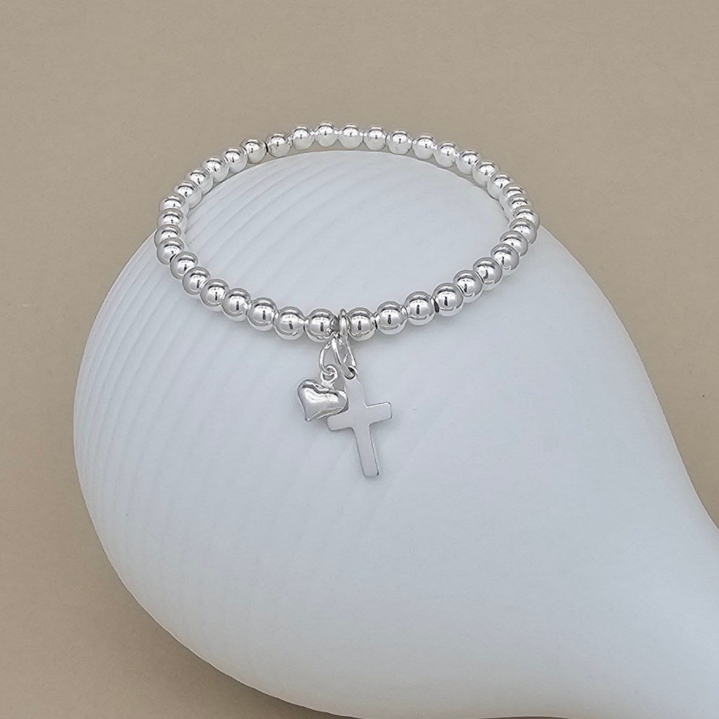 A silver beaded stretch bracelet with a small cross and heart charm, displayed on a white ceramic dish against a beige background.