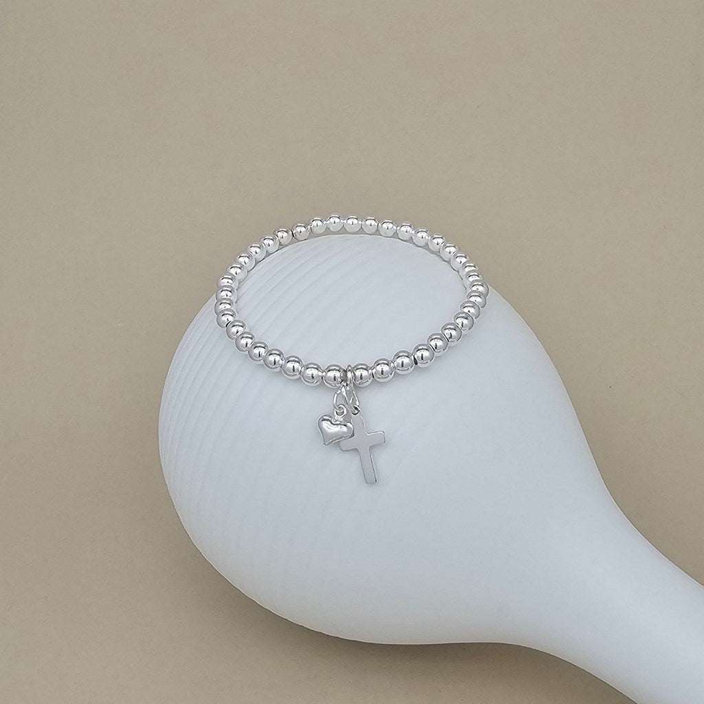 A silver beaded bracelet with a small cross and heart charm, displayed on a white ceramic stand against a warm beige background.