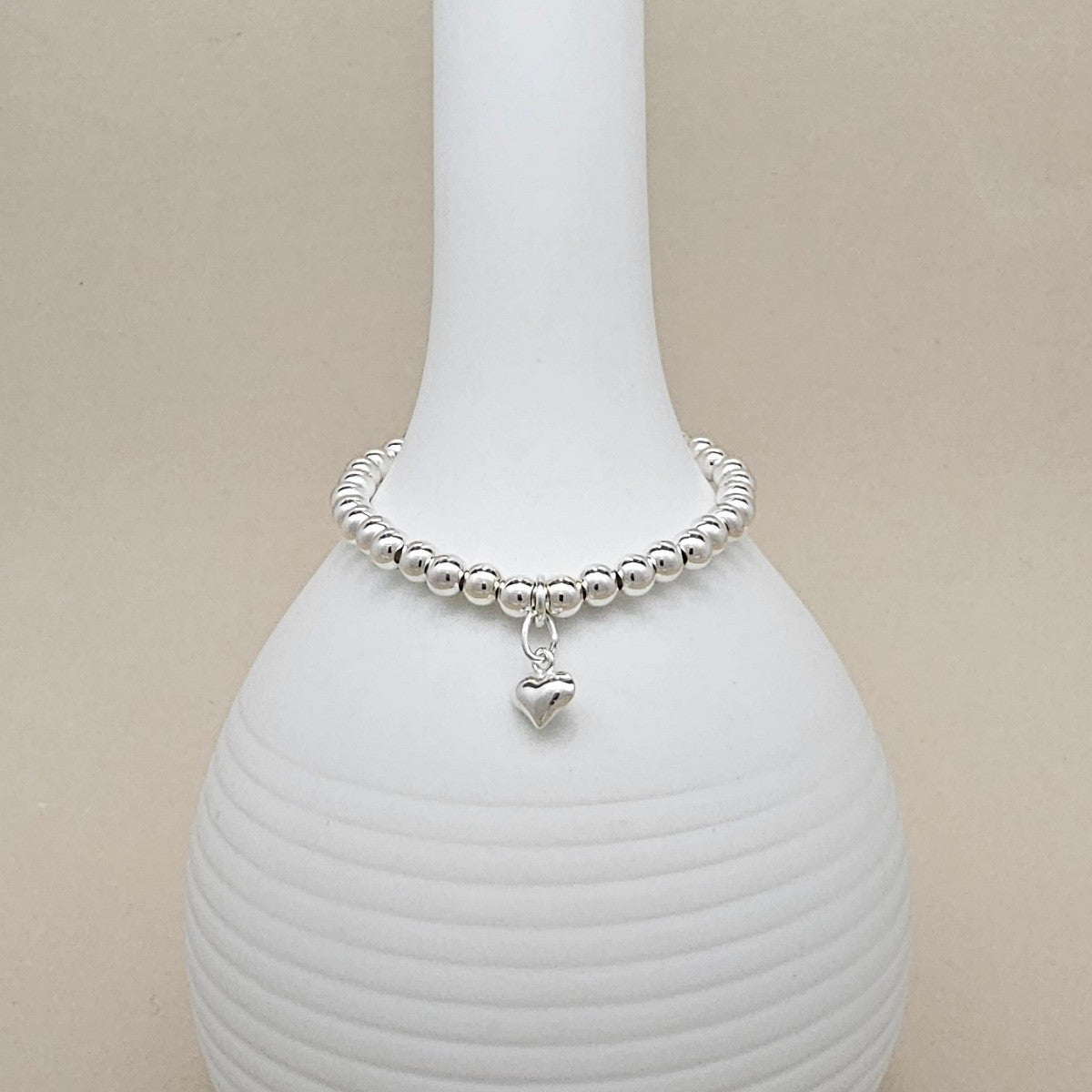 A silver beaded bracelet with a small dangling heart charm, displayed on a white ribbed ceramic vase against a warm beige background.