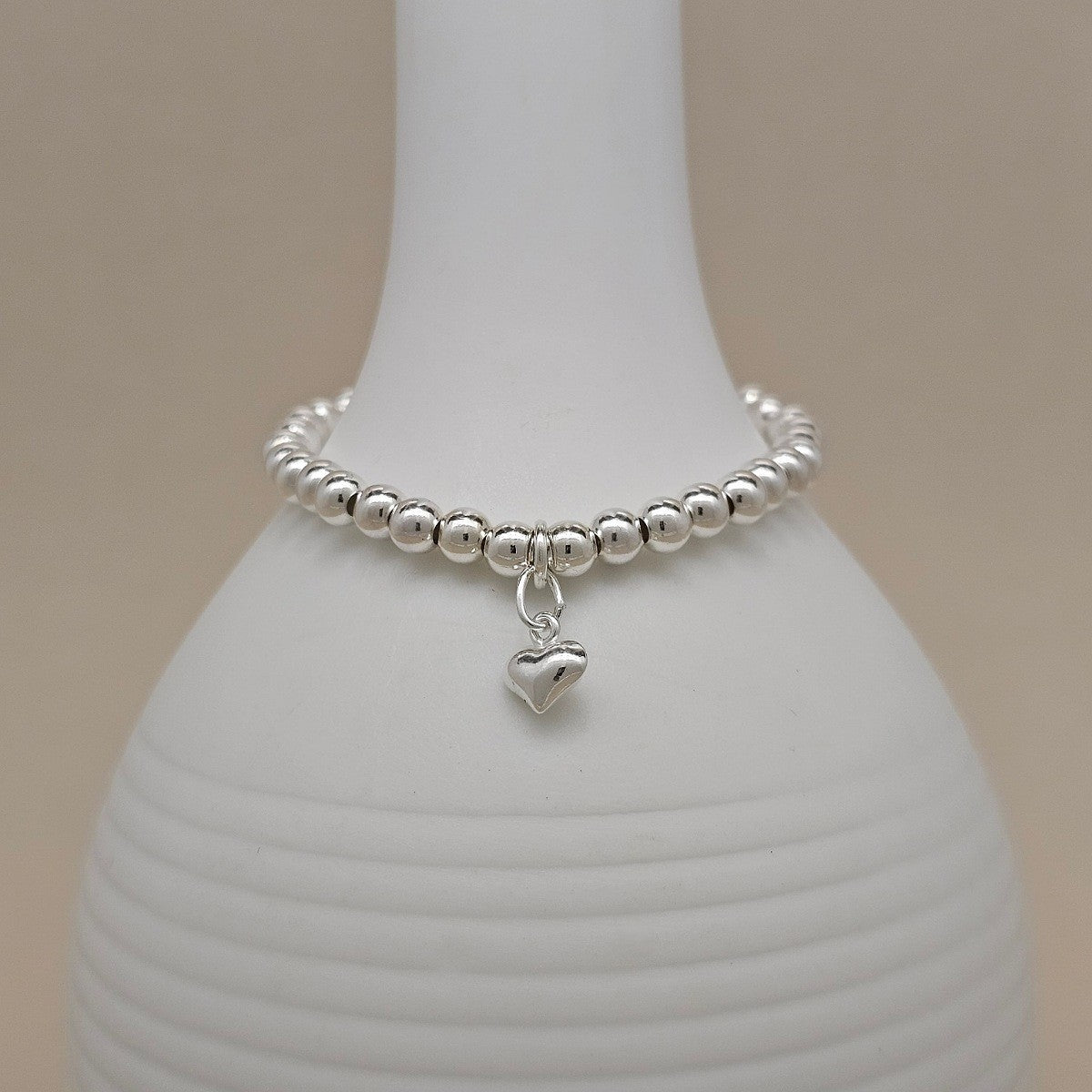 A silver beaded bracelet with a small dangling heart charm, displayed on a white ribbed ceramic vase against a warm beige background.