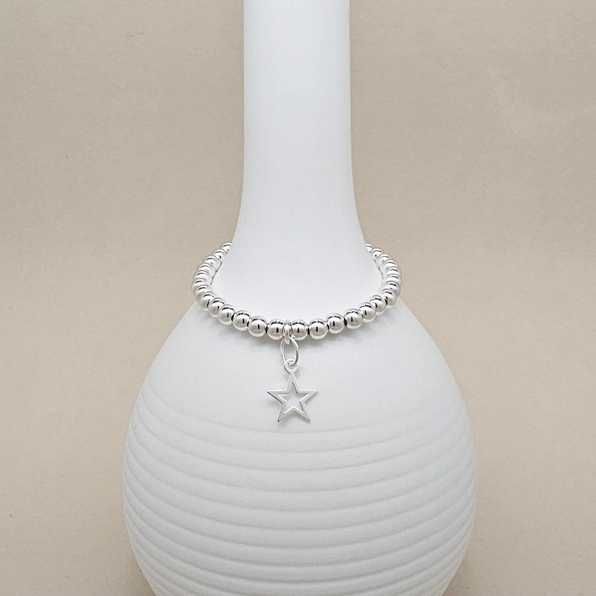 A silver beaded bracelet with a dangling star charm, displayed on a white ribbed ceramic vase against a beige background.