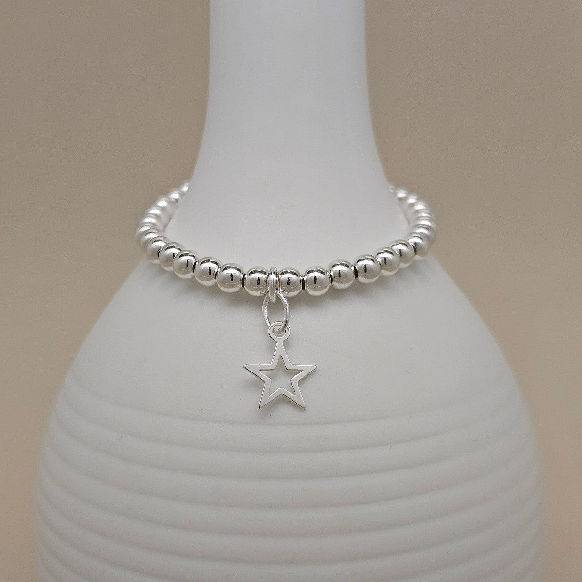 A silver beaded bracelet with a dangling open star charm, displayed on a white ceramic vase against a warm beige background.