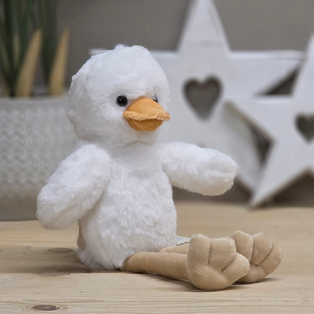 A white soft fuzzy plush chick with an orange beak and beige long legs, with black eyes.  On a wooden surface with a blurred star-shaped object in the background
