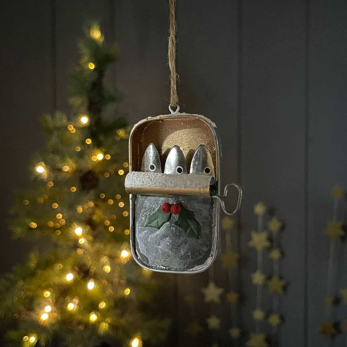 A decorative metal Christmas hanging ornament shaped like an open tin of sardines with three small fish faces and a holly accent, suspended by a twine string against a dark panelled background with blurred warm lights.