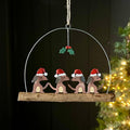 A hanging Christmas ornament shaped like four small brown dogs wearing red Santa hats, mounted on a rustic wooden branch and suspended from a metal arch with a holly accent, shown against a dark background with blurred holiday lights.