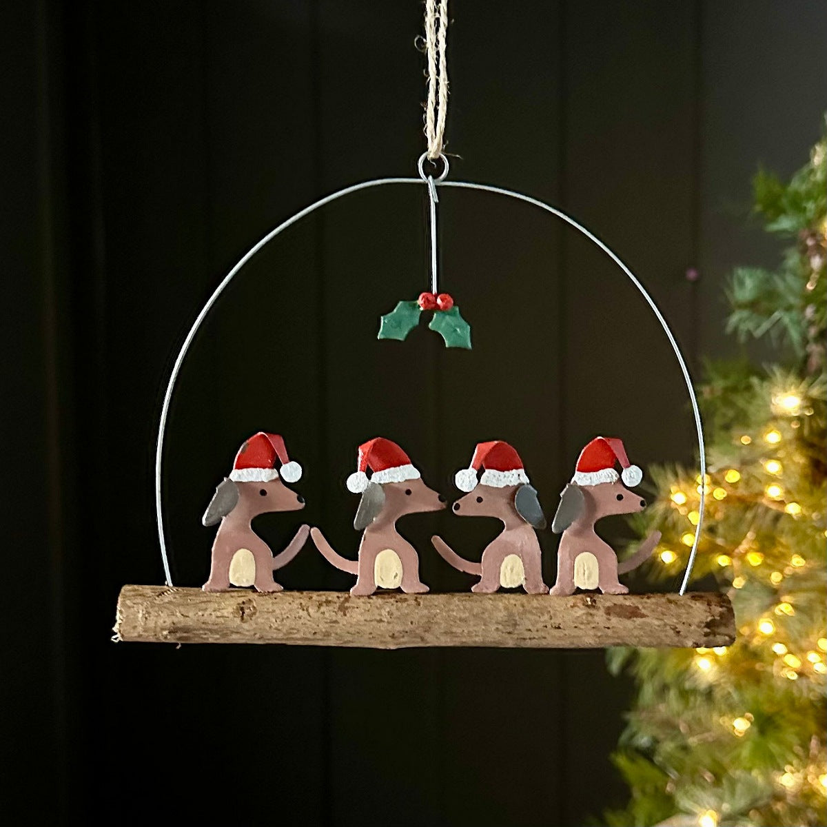 A hanging Christmas ornament shaped like four small brown dogs wearing red Santa hats, mounted on a rustic wooden branch and suspended from a metal arch with a holly accent, shown against a dark background with blurred holiday lights.