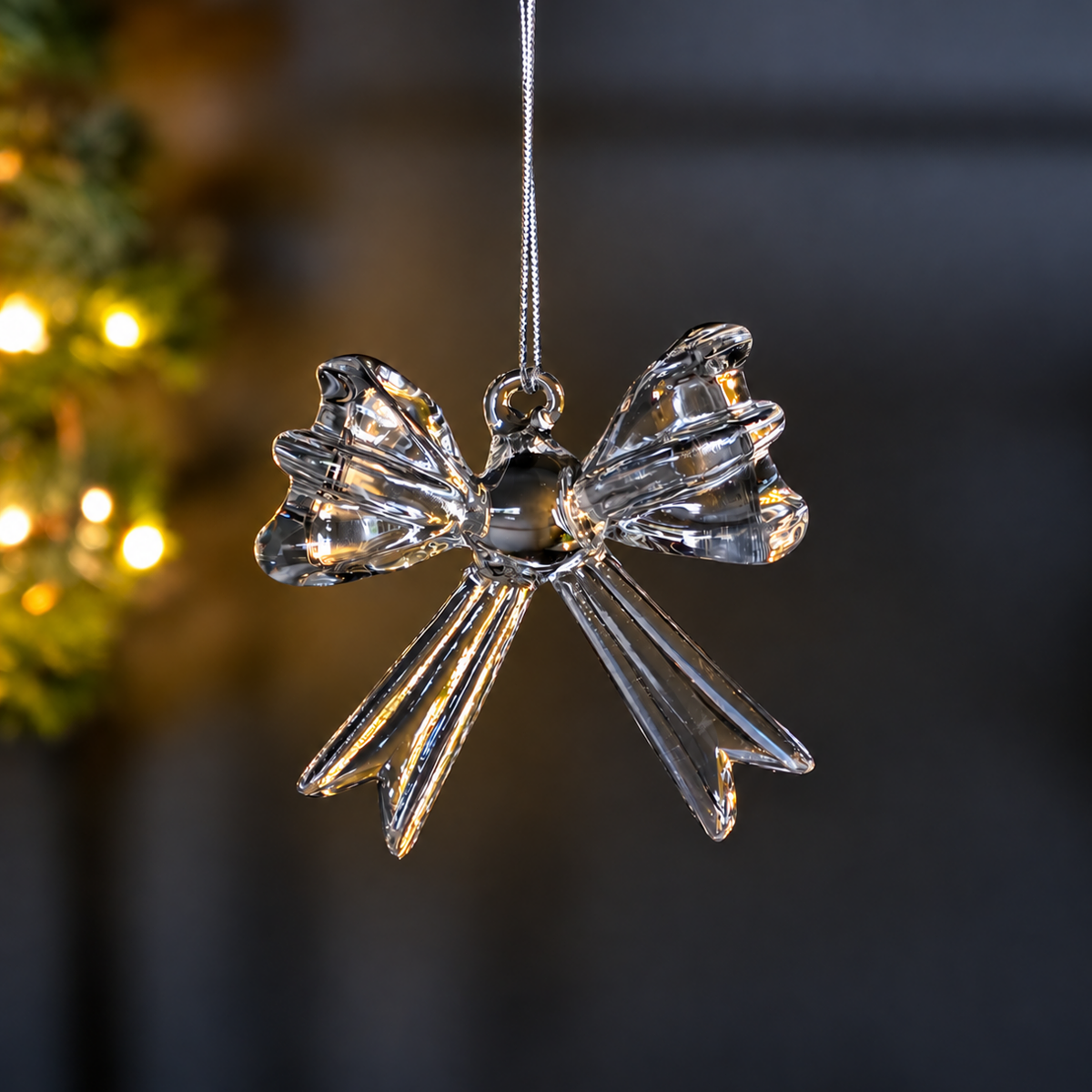 A clear hanging glass bow Christmas decoration hanging from a thin silver string against a dark, softly blurred background with warm bokeh lights on the left.