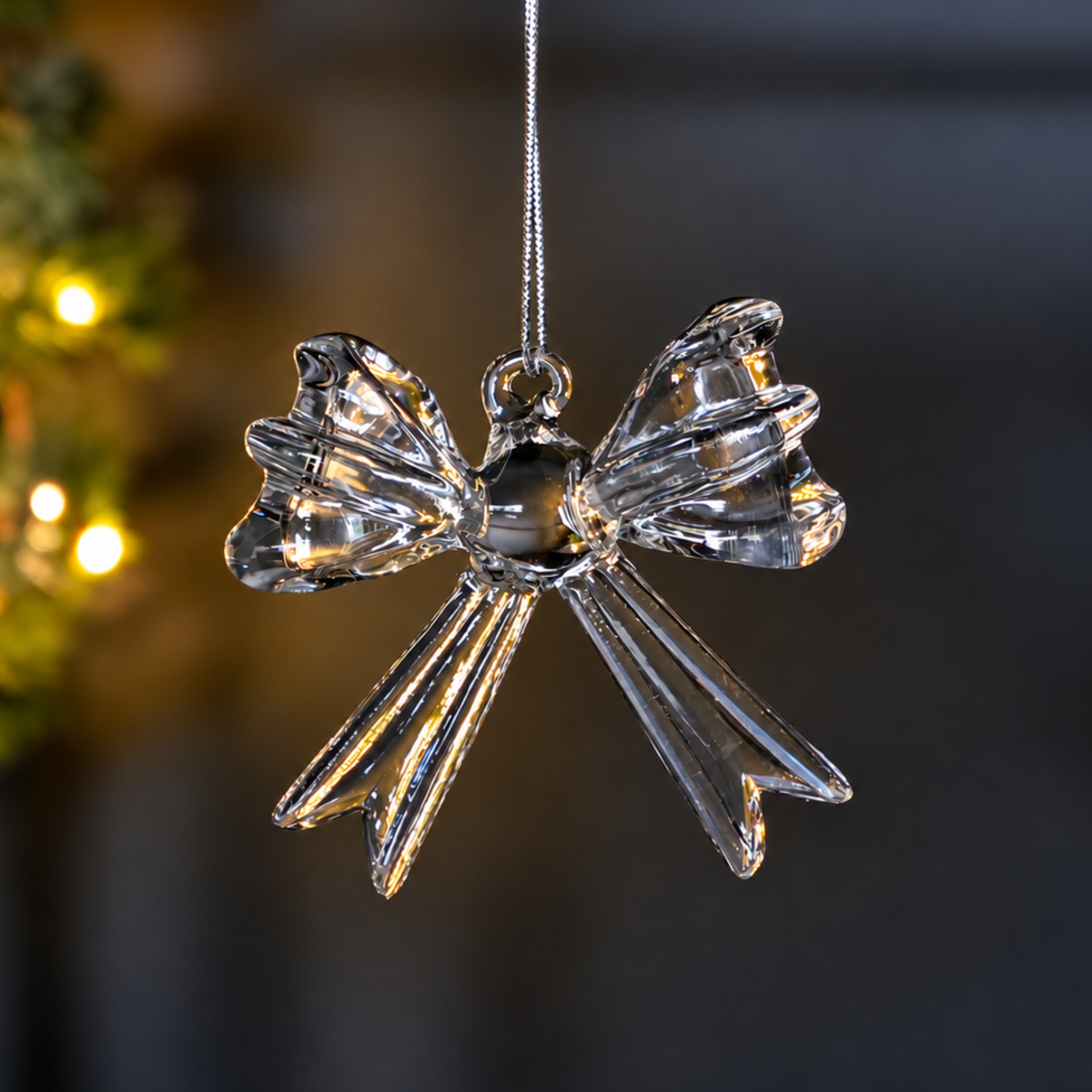 A clear glass bow Christmas ornament hanging from a thin silver string against a dark, softly blurred background with warm lights.