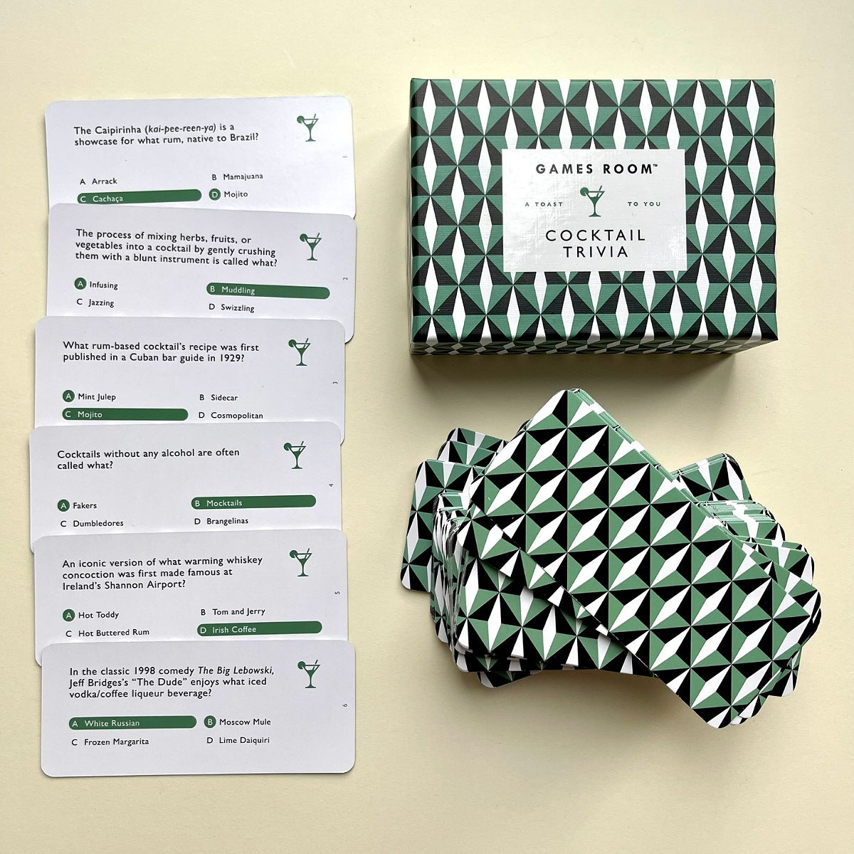 Games Room Cocktail Trivia set shown with a stack of white question cards, a green and white geometric patterned box, and several matching wrapped packages on a light beige background.