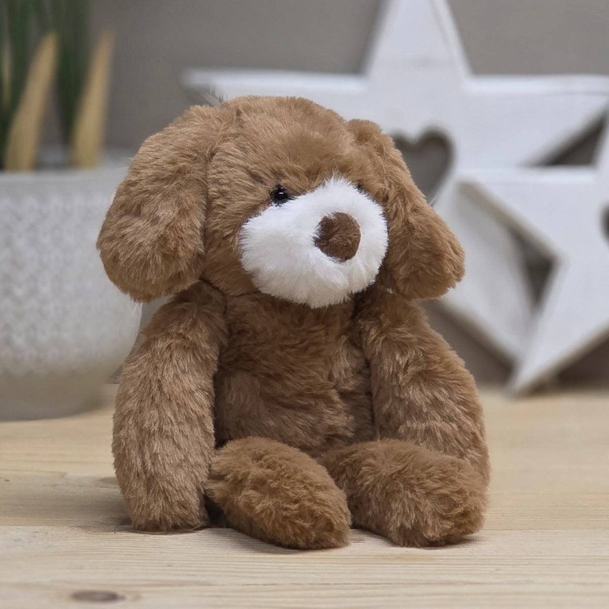A brown plush puppy toy with a white snout and black eyes, sitting on a light wooden surface with a blurred decorative background.
