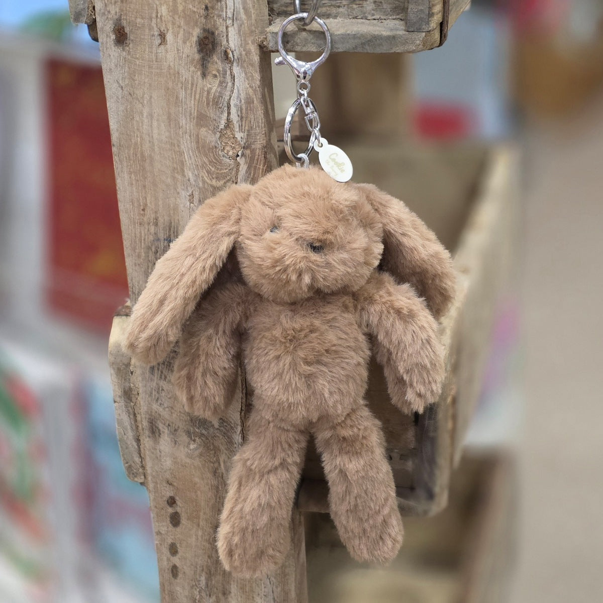 A brown plush bunny keychain hanging from a rustic wooden post. The keychain features a soft, fuzzy brown bunny with black eyes and nose, attached to a silver metal key ring and a small round tag.

