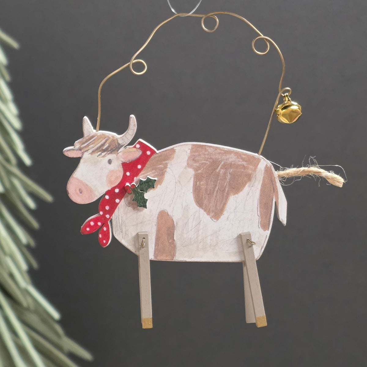 Hand-painted wooden cow Christmas ornament with brown and white patches, a red polka-dot scarf, and a small gold hanging bell, shown against a dark grey background with pine needles at the side.