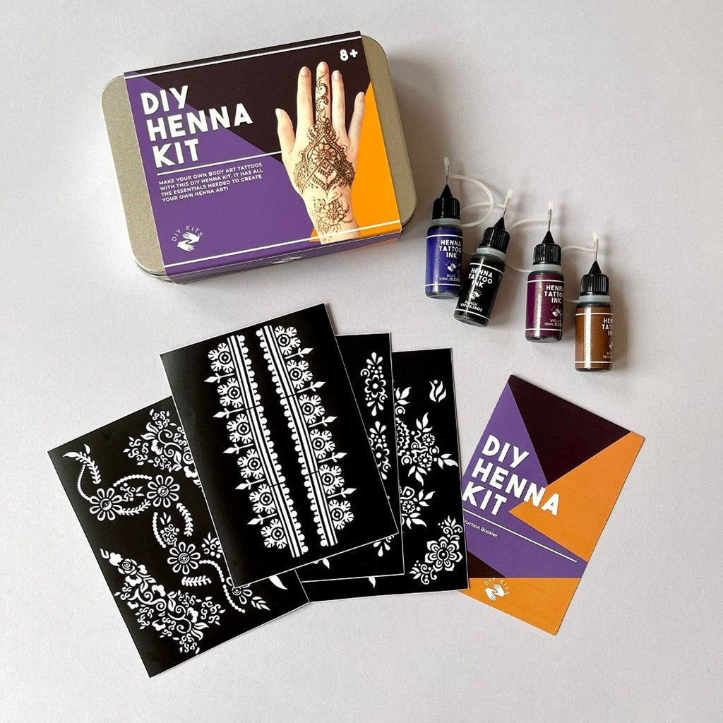 DIY Henna Kit set laid out on a light grey surface, including a metal tin with a henna kit label, several black henna stencil sheets with white floral patterns, four small dye bottles in purple, brown, and dark tones, and a small instruction card.