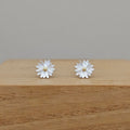 A pair of silver daisy stud earrings with textured petals and small gold centers, displayed on a wooden block against a neutral gray background.