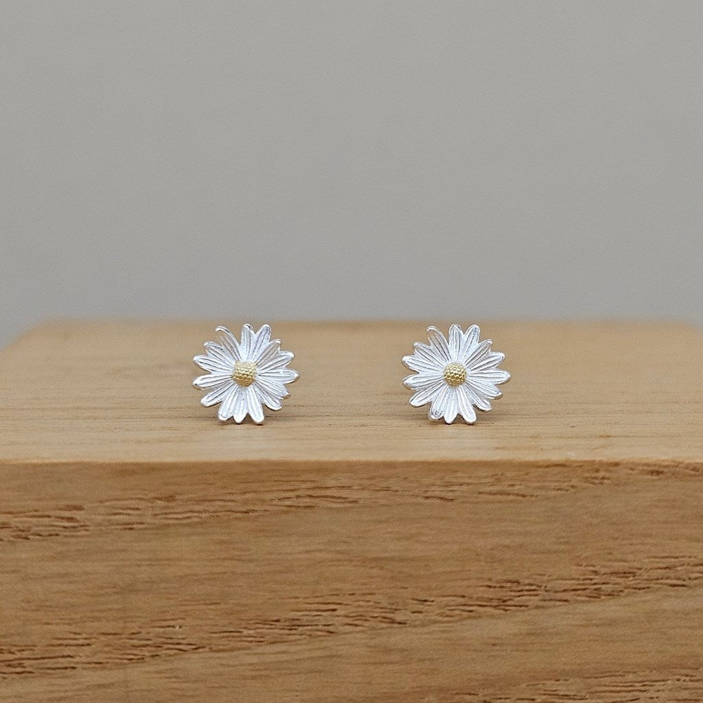 A pair of silver daisy stud earrings with textured petals and small gold centers, displayed on a wooden block against a neutral gray background.