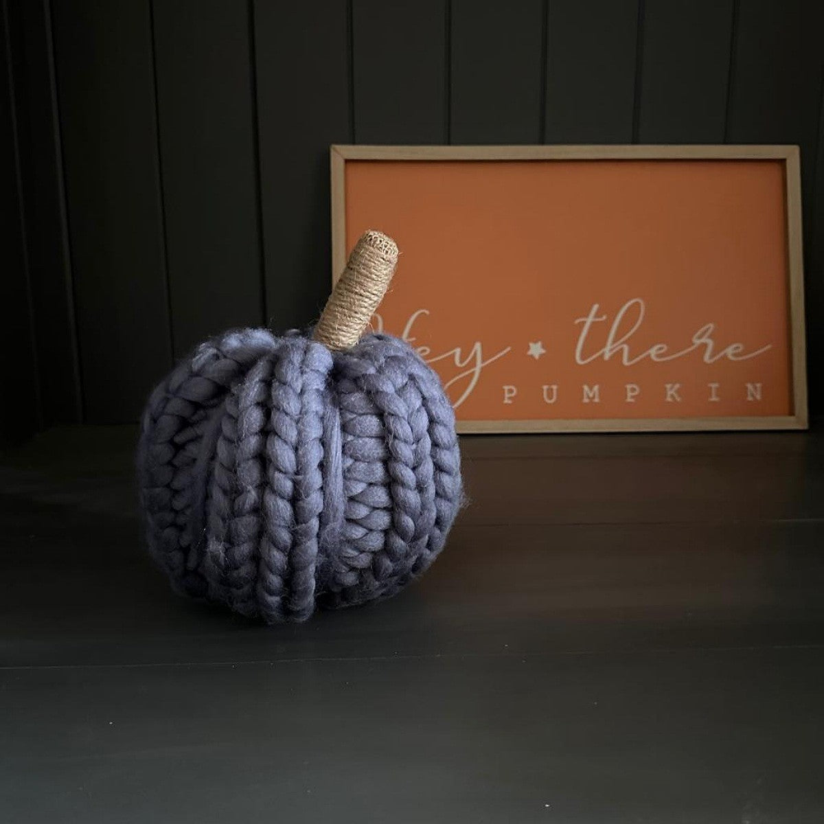 Knitted wooden dark grey pumpkin decoration on a dark surface with a framed sign in the background.