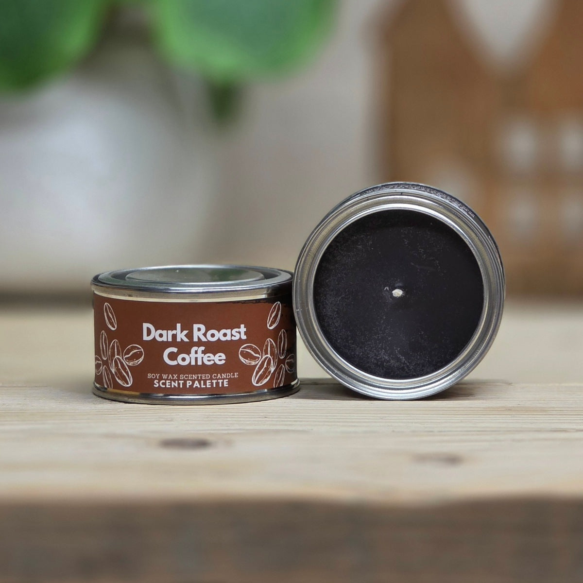 A small round tin candle labeled 'Dark Roast Coffee' sits on a wooden surface with its lid removed and placed beside it, showing a dark coffee-brown wax inside. The background is softly blurred with green leaves and a warm indoor setting.
