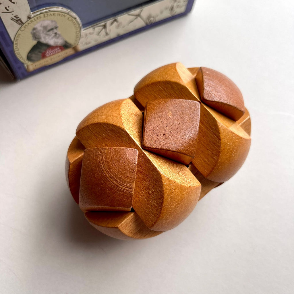 A wooden puzzle box with a complex interlocking geometric design, shown on a light grey surface with its blue and white packaging box partially visible in the background.