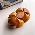 A wooden puzzle box with a complex interlocking geometric design, shown on a light grey surface with its blue and white packaging box partially visible in the background.