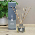 Deep Sea Minerals reed diffuser set on a light wood surface, featuring a clear glass bottle with natural reeds and its blue-gray packaging box next to a potted green plant.