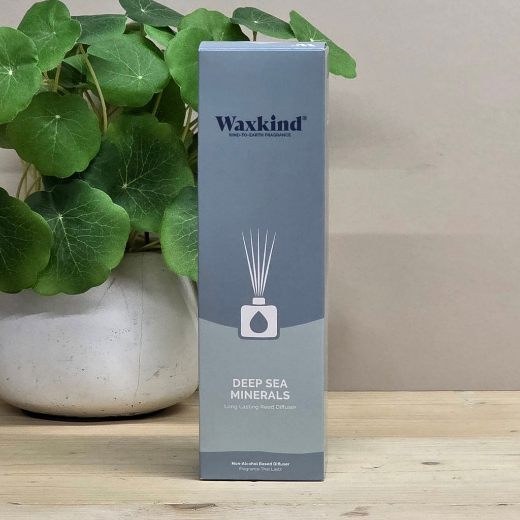 A tall rectangular Waxkind reed diffuser box labeled 'Deep Sea Minerals' stands on a light wood surface beside a potted green plant against a beige wall.