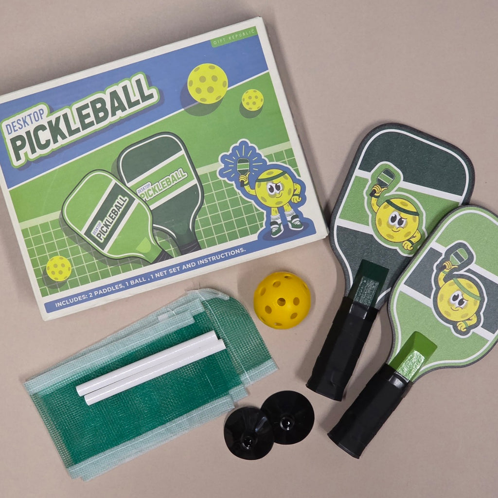 A complete set of desk pickleball toys arranged on a neutral beige background, including a product box, two paddle-shaped items, a yellow ball, a mesh case with white paddles, and two black discs.