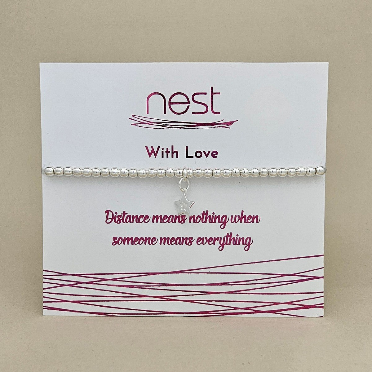 A silver beaded bracelet with a small star charm displayed on a white card from the brand Nest. The card features the text 'With Love' and 'Distance means nothing when someone means everything' in dark pink lettering, set against a neutral beige background.