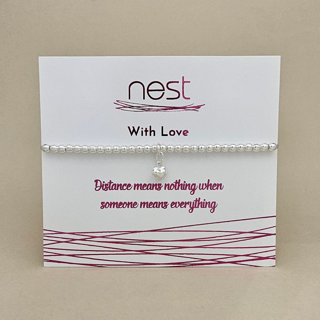 A silver beaded bracelet with a small heart-shaped crystal charm, displayed on a white card from the brand 'nest' with the message 'With Love' and 'Distance means nothing when someone means everything'.
