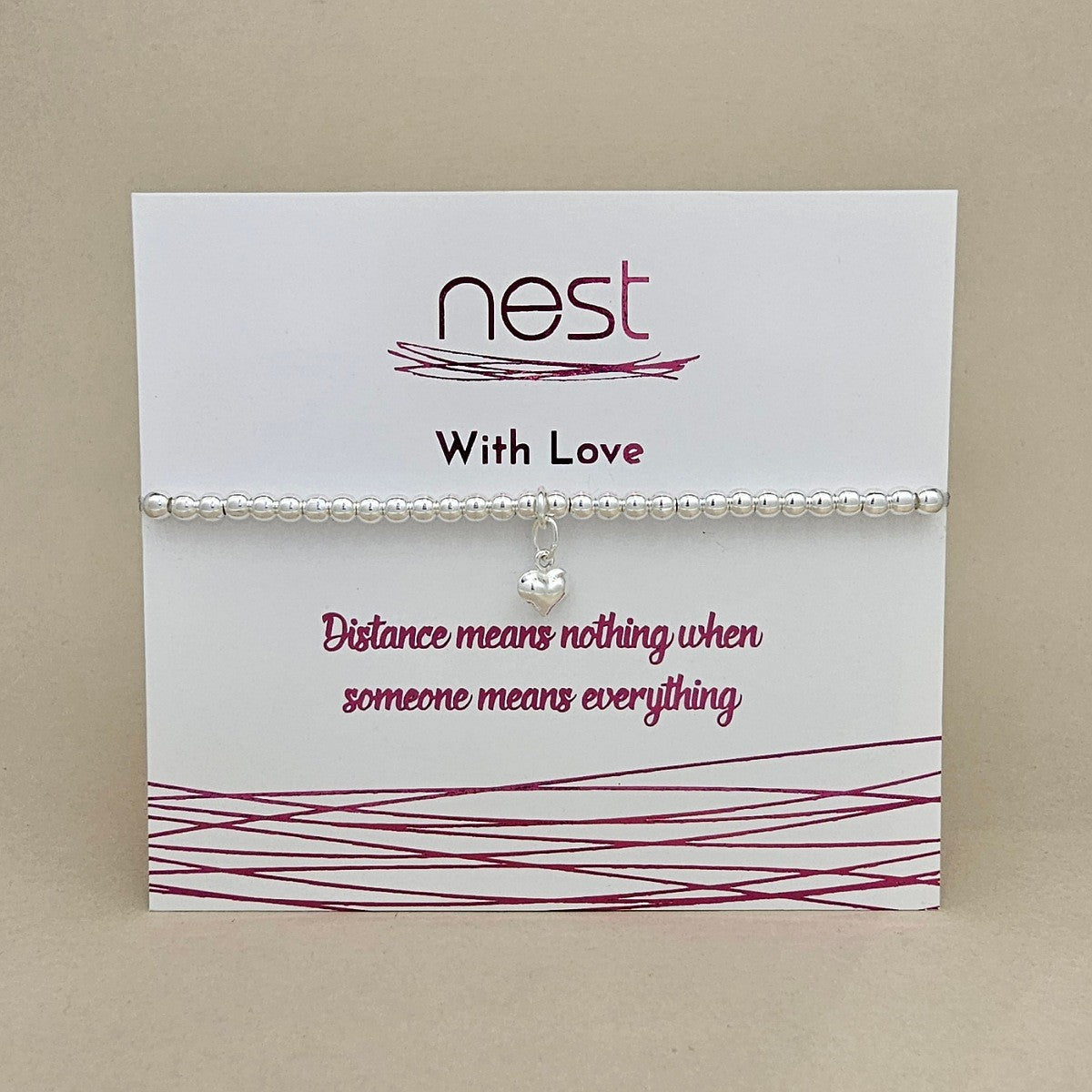 A silver beaded bracelet with a small heart-shaped crystal charm, displayed on a white card from the brand 'nest' with the message 'With Love' and 'Distance means nothing when someone means everything'.