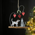 A rustic metal hanging ornament with two dogs sitting together on a wooden branch, with three red heart ornaments and mistletoe hanging above, photographed against a dark background with blurred Christmas tree lights at the side.