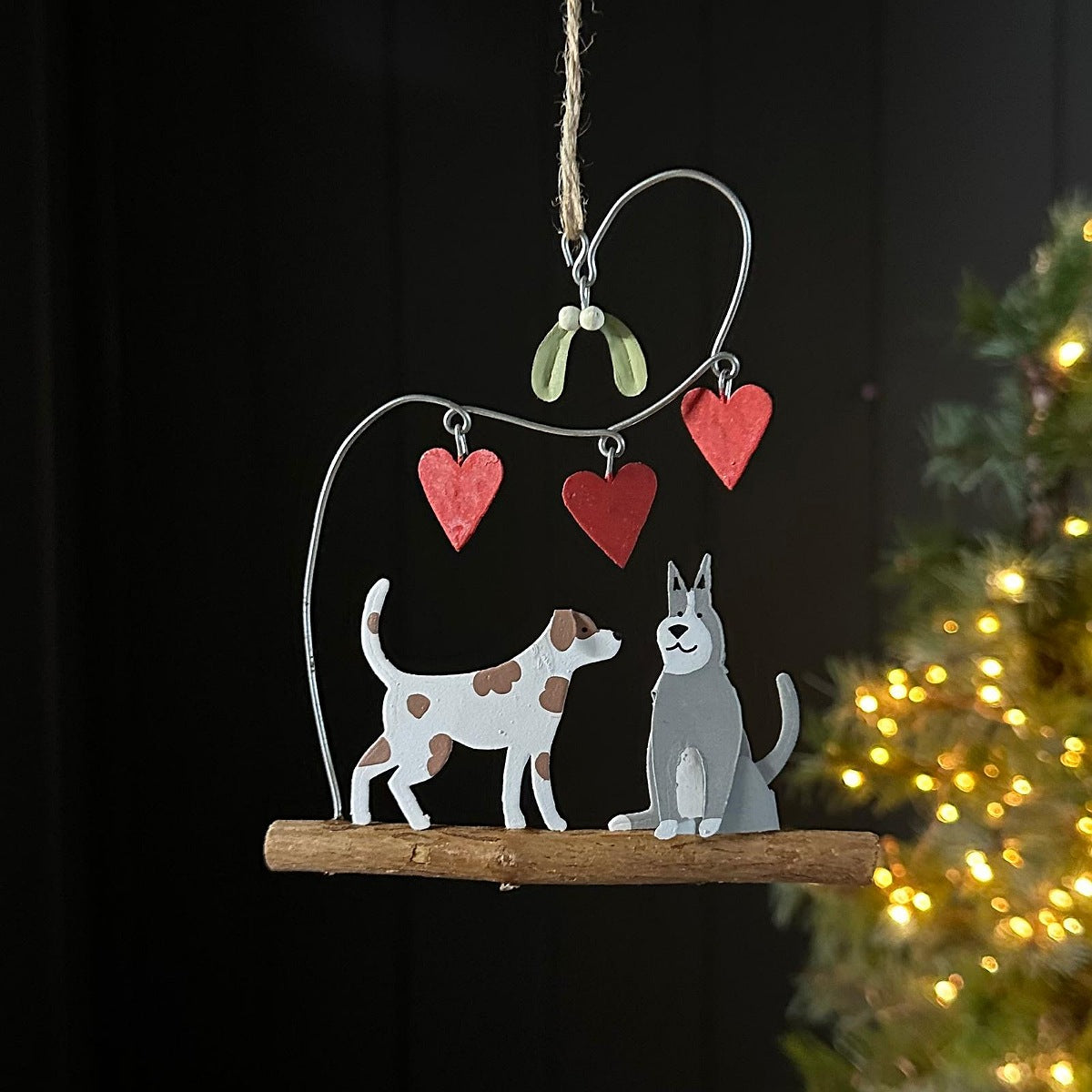 A rustic metal hanging ornament with two dogs sitting together on a wooden branch, with three red heart ornaments and mistletoe hanging above, photographed against a dark background with blurred Christmas tree lights at the side.