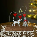 A festive metal Christmas ornament featuring two dogs under a wire heart arch with red hearts, mistletoe, and a red bow, displayed on a wooden base surrounded by white decorative beads with a blurred Christmas tree in the background.