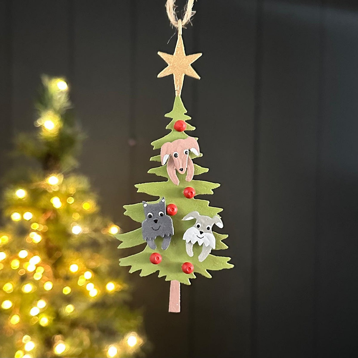 A hanging metal Christmas decoration of a green tree with a gold star with dog figures and red bauble decorations, photographed in front of a softly blurred lit Christmas tree.