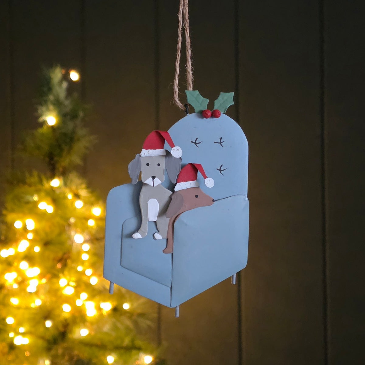 A festive metal hanging decoration of two dogs with Santa hats sat on a light grey chair with blurred Christmas light tree behind.