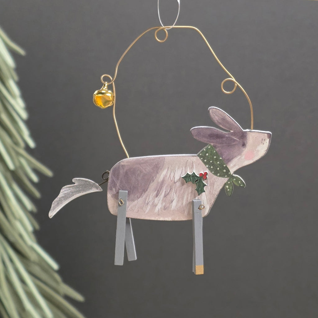Handcrafted wooden Christmas dog hanging ornament, with a gold wire hoop, small amber bell, green bow, and gry legs, shown against a dark grey background with blurred pine needles at the side.