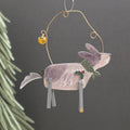 Handcrafted wooden Christmas dog hanging ornament, with a gold wire hoop, small amber bell, green bow, and gry legs, shown against a dark grey background with blurred pine needles at the side.