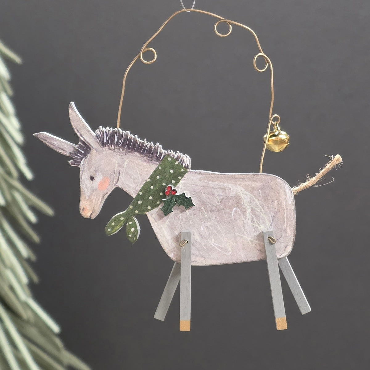 A handcrafted wooden donkey Christmas ornament with a pale pinkish-grey body, green and white patterned scarf, small red holly accents, and a gold hanging wire frame with a small gold bell.