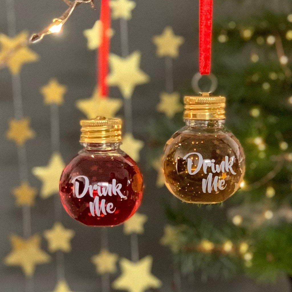 Two small plastic hanging Christmas ornaments shaped like glass bottles with gold caps and red ribbons. One ornament is filled with red liquid and the other with amber liquid, both printed with the phrase 'Drink Me' in white script.