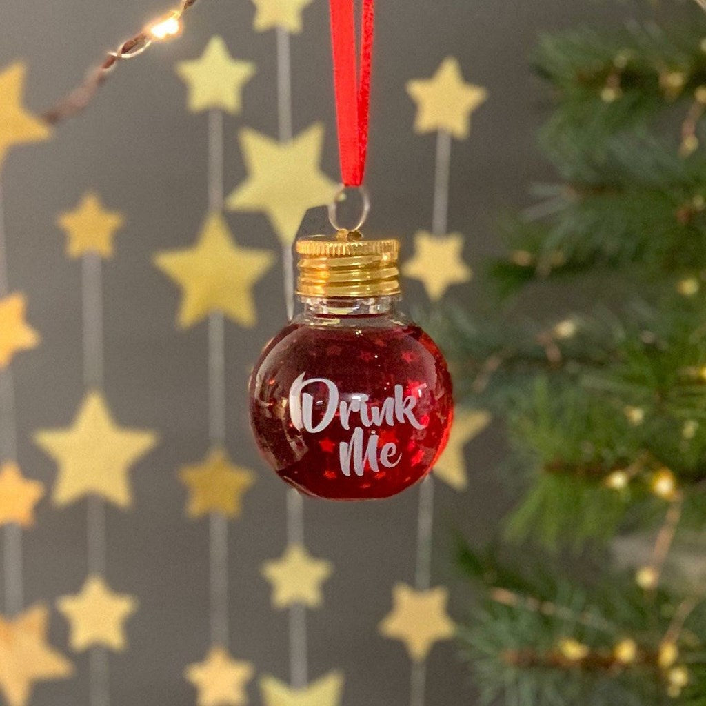 Plastic ornament with 'Drink Me' text, gold cap, on a string against a festive background with stars and Christmas tree.
