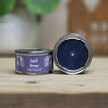 A small round metal candle tin labeled 'Earl Grey' sits on a wooden surface with its lid removed and placed beside it, showing a dark blue wax surface inside. The background is softly blurred with green leaves and warm indoor decor.