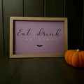 A frame with the words 'Eat, drink + BE SCARY' in black and white lettering on a dusty purple background, set in a light wood frame and styled on a dark wooden surface next to an orange pumpkin.