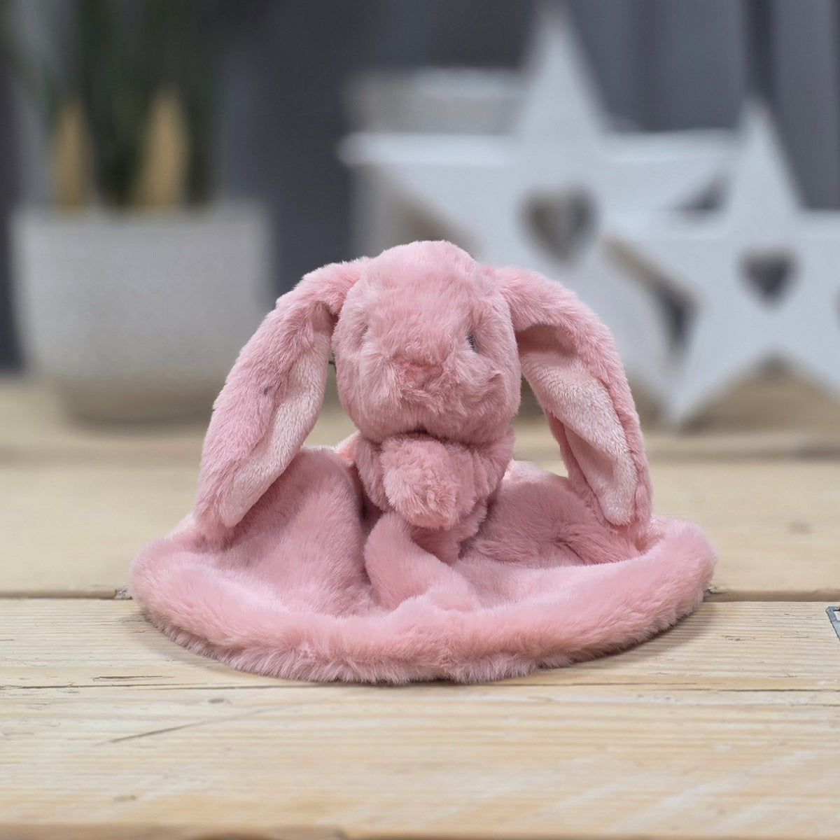 A pink plush bunny head blanket with long floppy ears sits on a light wood surface, with a softly blurred home decor background featuring a white star-shaped ornament and a potted plant.