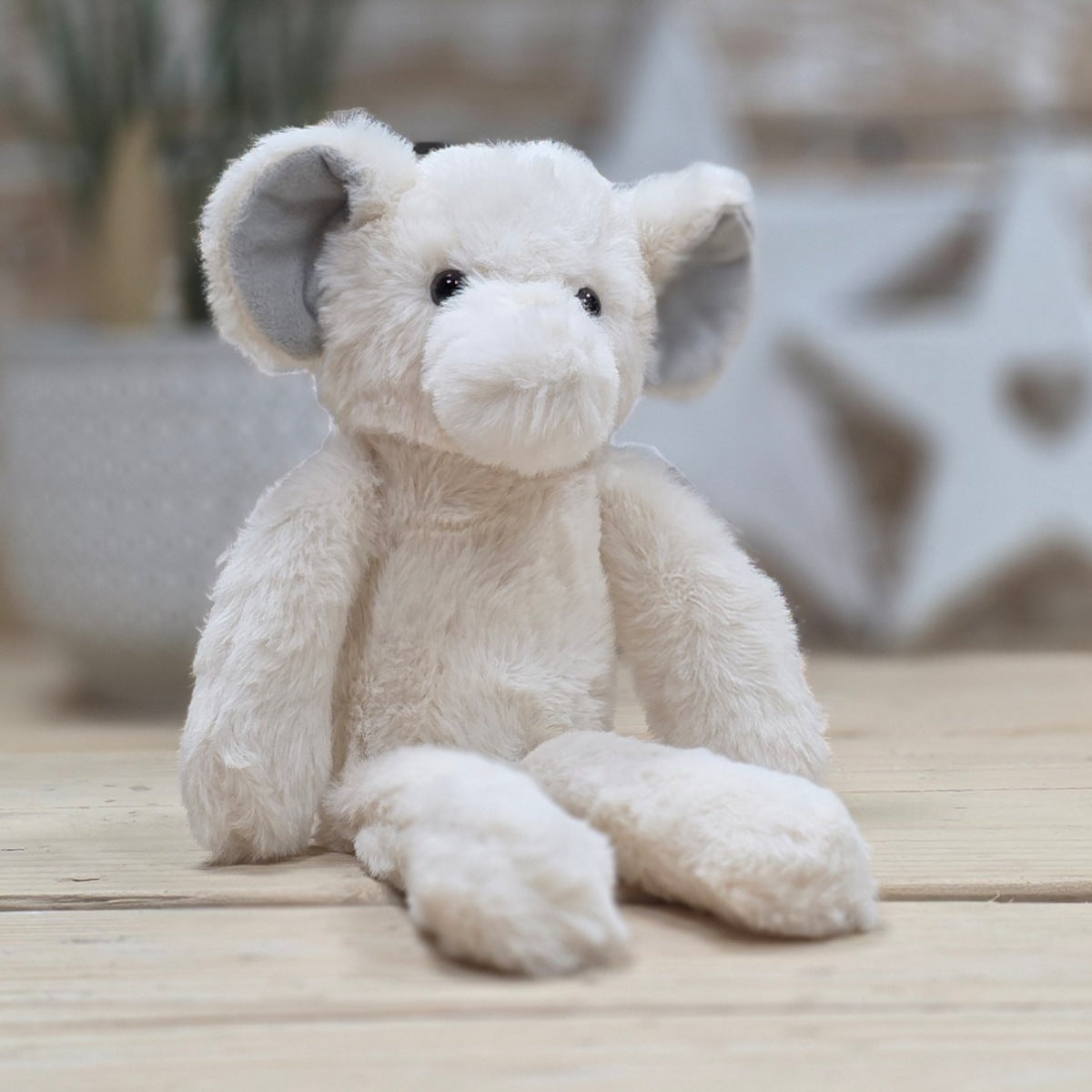 Cream plush soft fuzzy elephant toy  with grey ears and black eyes, on a wooden surface with a blurred natural background
