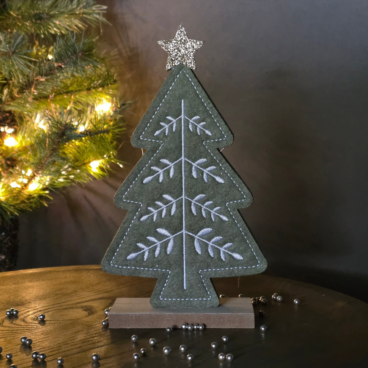 A felt Christmas tree tabletop decoration in muted green with white embroidered branches and a silver star topper, displayed on a wooden stand on a table with a blurred lit Christmas tree in the background.