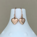 A pair of rose gold heart-shaped drop earrings with curved hooks, displayed on a white ceramic jewelry bust against a neutral beige background.