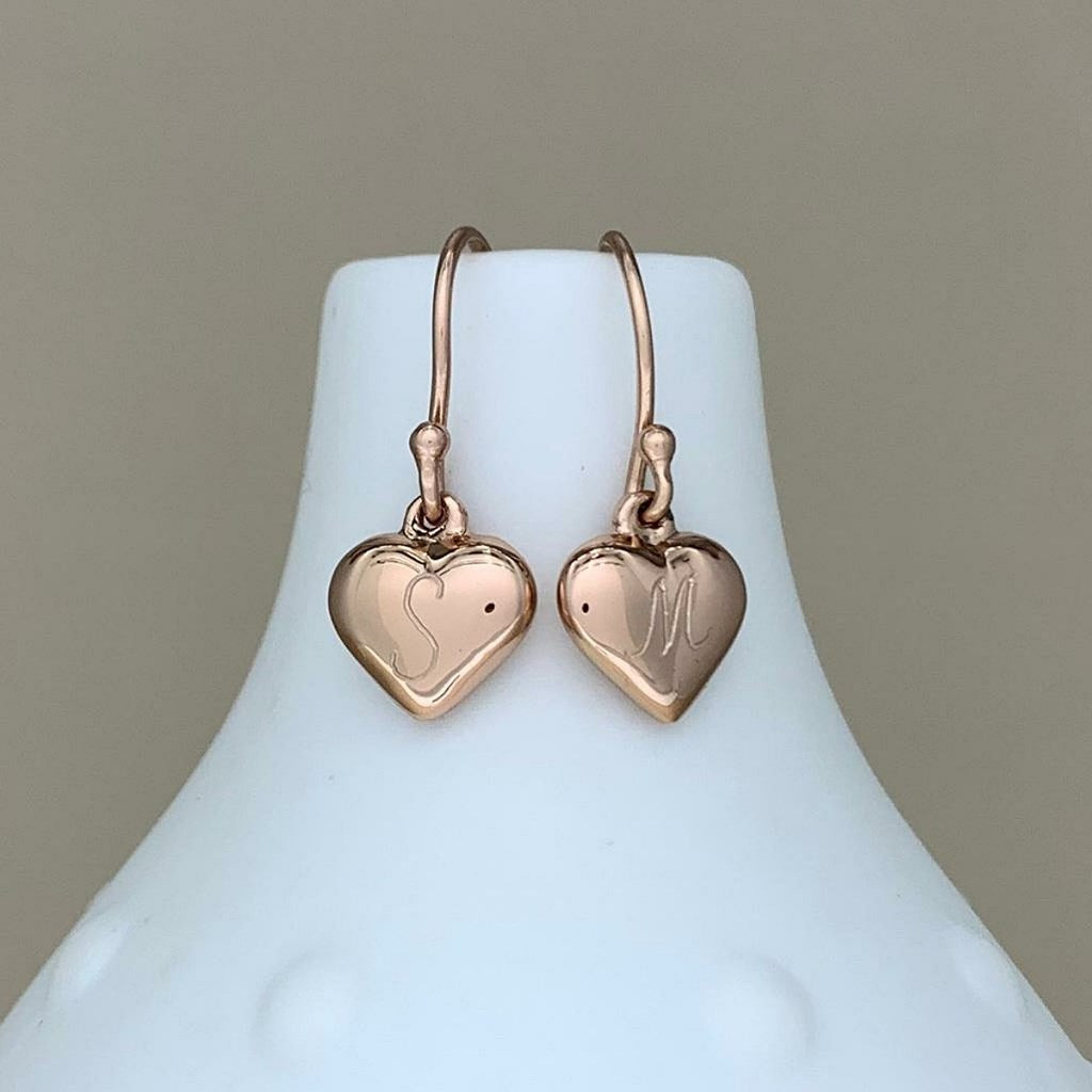 A pair of rose gold heart-shaped drop earrings with curved hooks, displayed on a white ceramic jewelry bust against a neutral beige background.