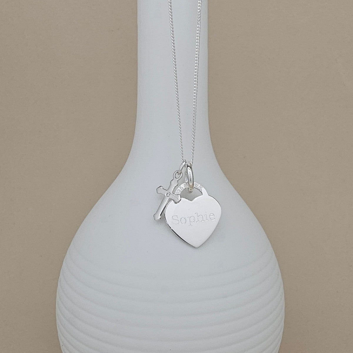 A silver necklace with a heart-shaped pendant engraved with the name 'Sophie' and a small cross charm, displayed on a ribbed white vase against a beige background.