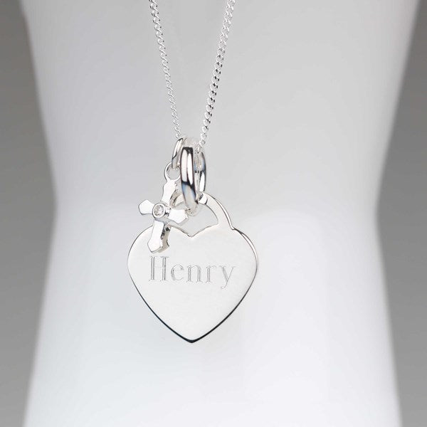 A silver heart-shaped pendant necklace engraved with the name 'Henry' and a small hanging cross charm, displayed on a white mannequin bust against a soft gray background.