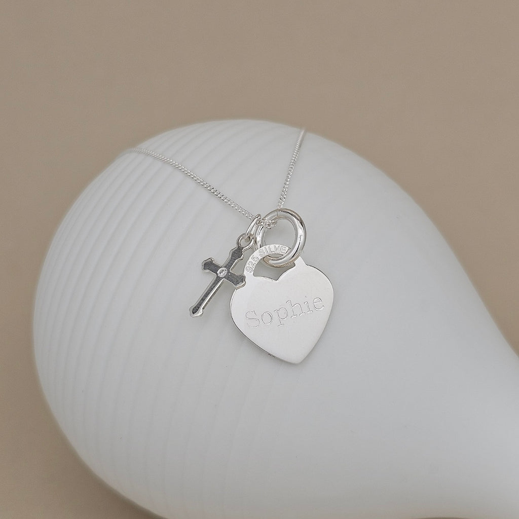 A silver necklace with a heart-shaped pendant engraved with the name 'Sophie' and a small cross charm, displayed on a white ribbed ceramic stand against a beige background.